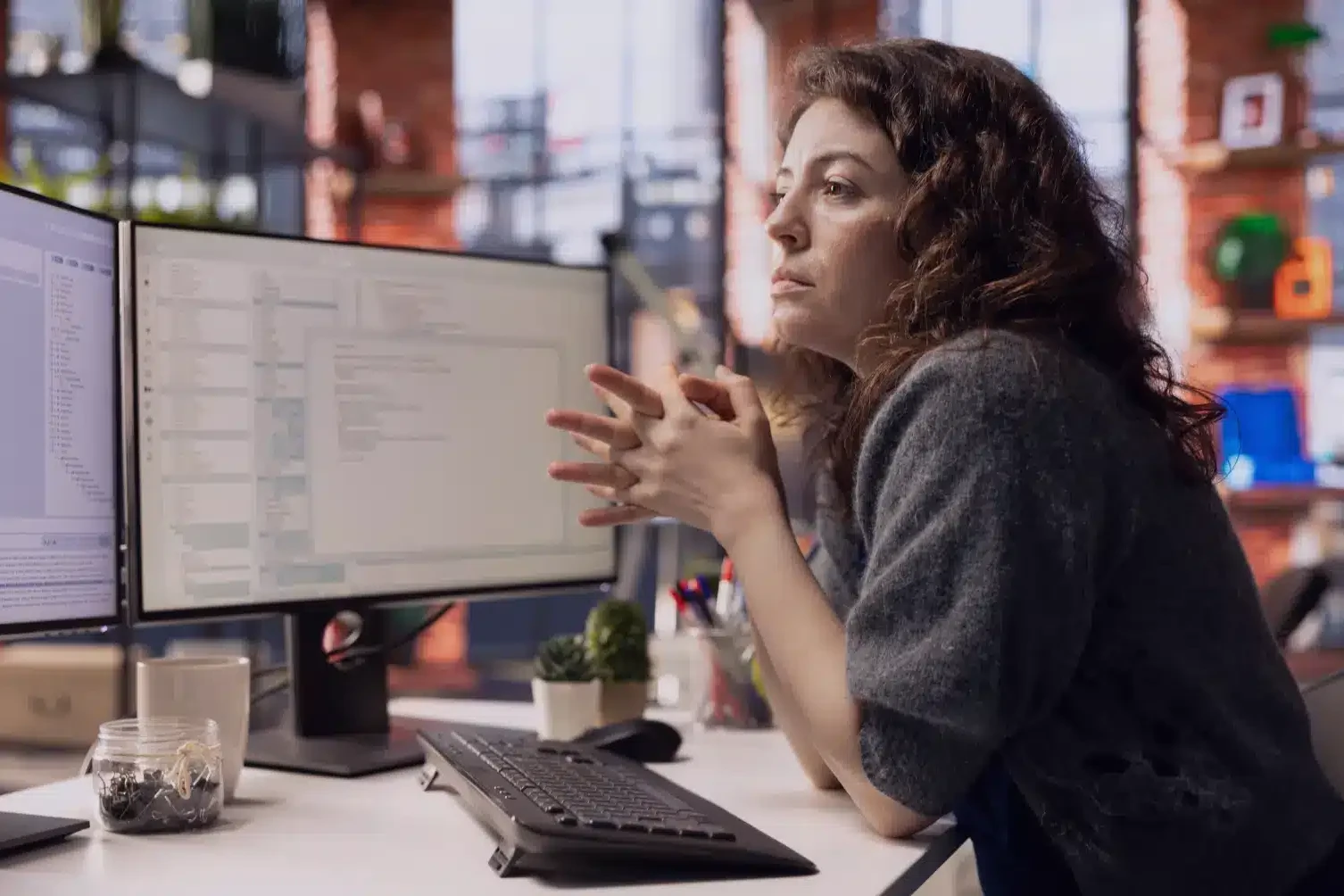 Woman at a desk with two computer monitors, gesturing as she explains something in a modern office.
